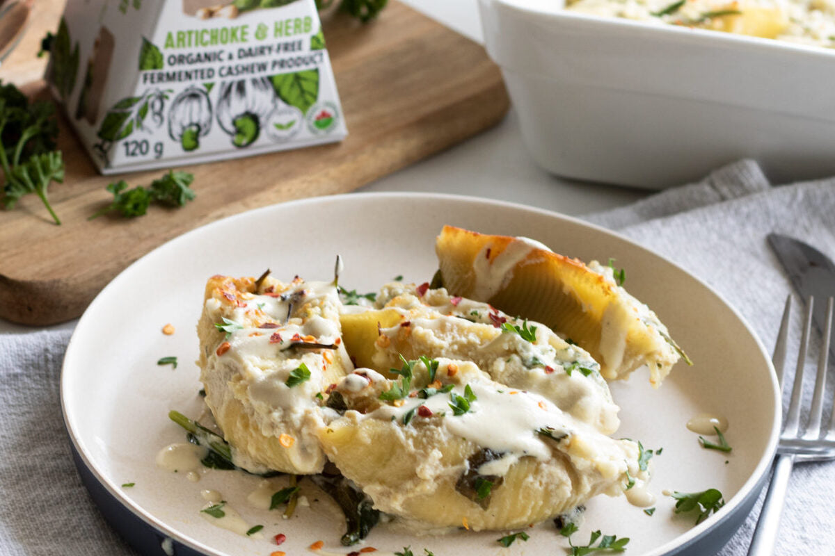 Assiette avec de grandes coquilles de pâtes farcies de fromage sans lait et garnies d'herbes fraîches. Placée à côté d'une boîte de fromage sans lait aux artichauts et aux herbes.