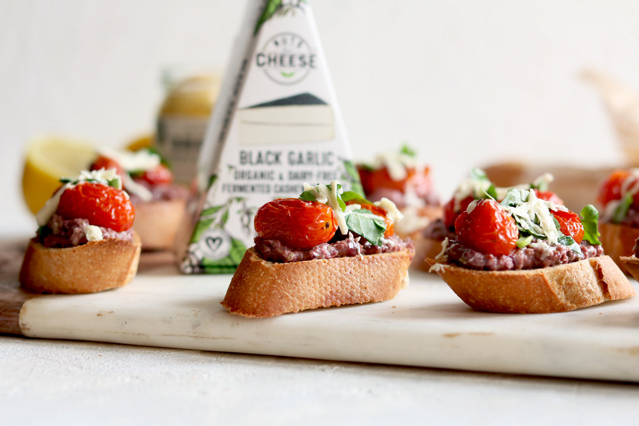 Trois tranches de baguette garnies de tapenade d'olives et de fromage à l'ail noir sans produits laitiers, de tomates cerises rôties et d'herbes fraîches. Servi à côté d'une boîte de fromage à l'ail noir sans produits laitiers.