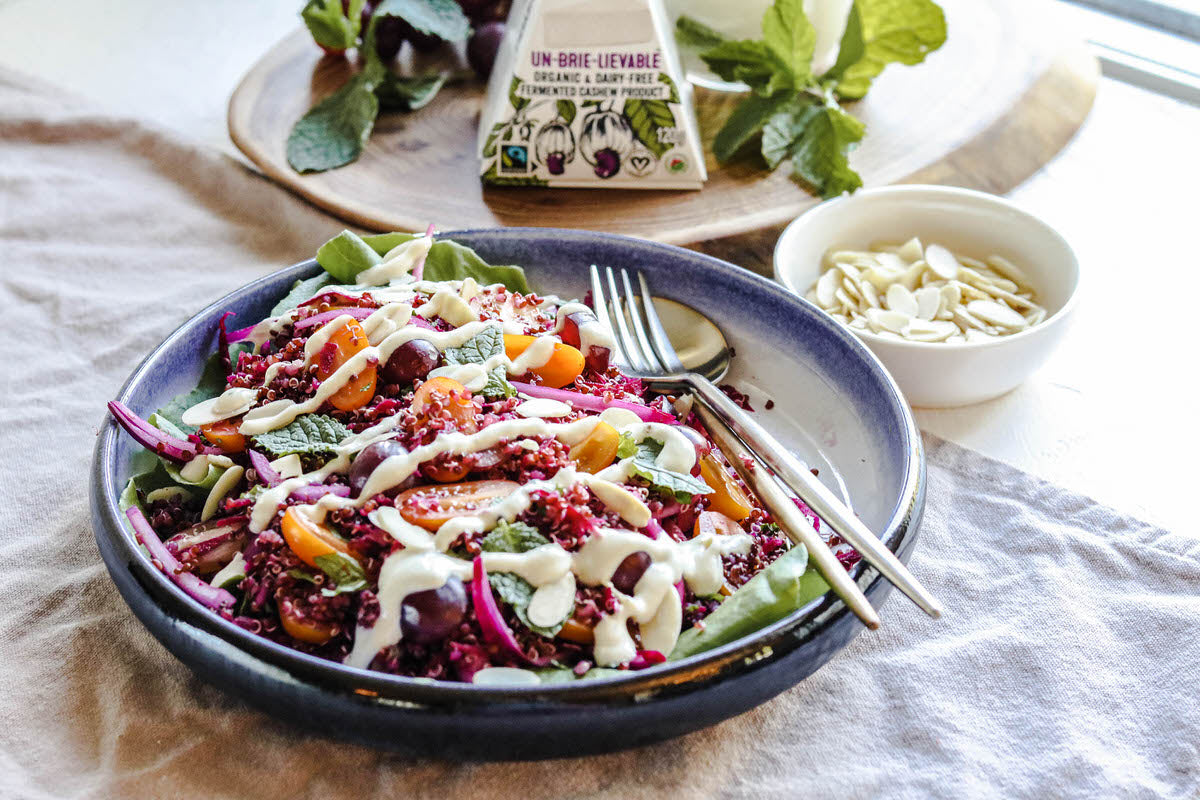 Bol de salade garni de quinoa rouge, de tomates fraîches, d'oignons rouges et d'un filet de vinaigrette sans produits laitiers. Servi à côté d'un bol d'amandes effilées et d'une boîte de fromage brie sans lait.