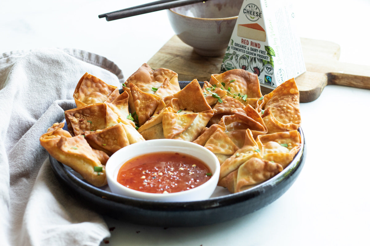 Grand bol de wontons végétaliens frits à l'air, farcis de gouda sans lait et servis avec un bol de sauce chili sucrée. Boîte de gouda fumé sans produits laitiers en arrière-plan.
