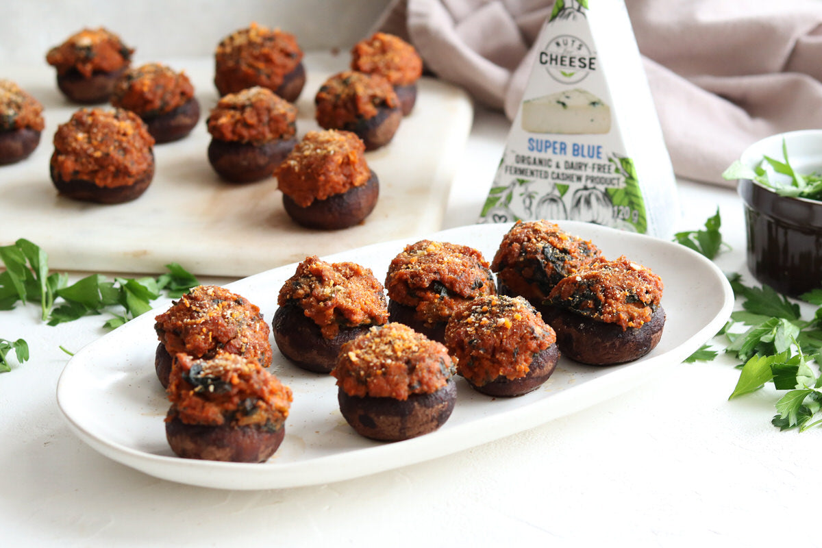 Deux assiettes de chapeaux de champignons cuits au four, farcis de saucisse chorizo végétalienne et de fromage bleu sans produits laitiers. Servis à côté d'une boîte de fromage bleu sans produits laitiers.