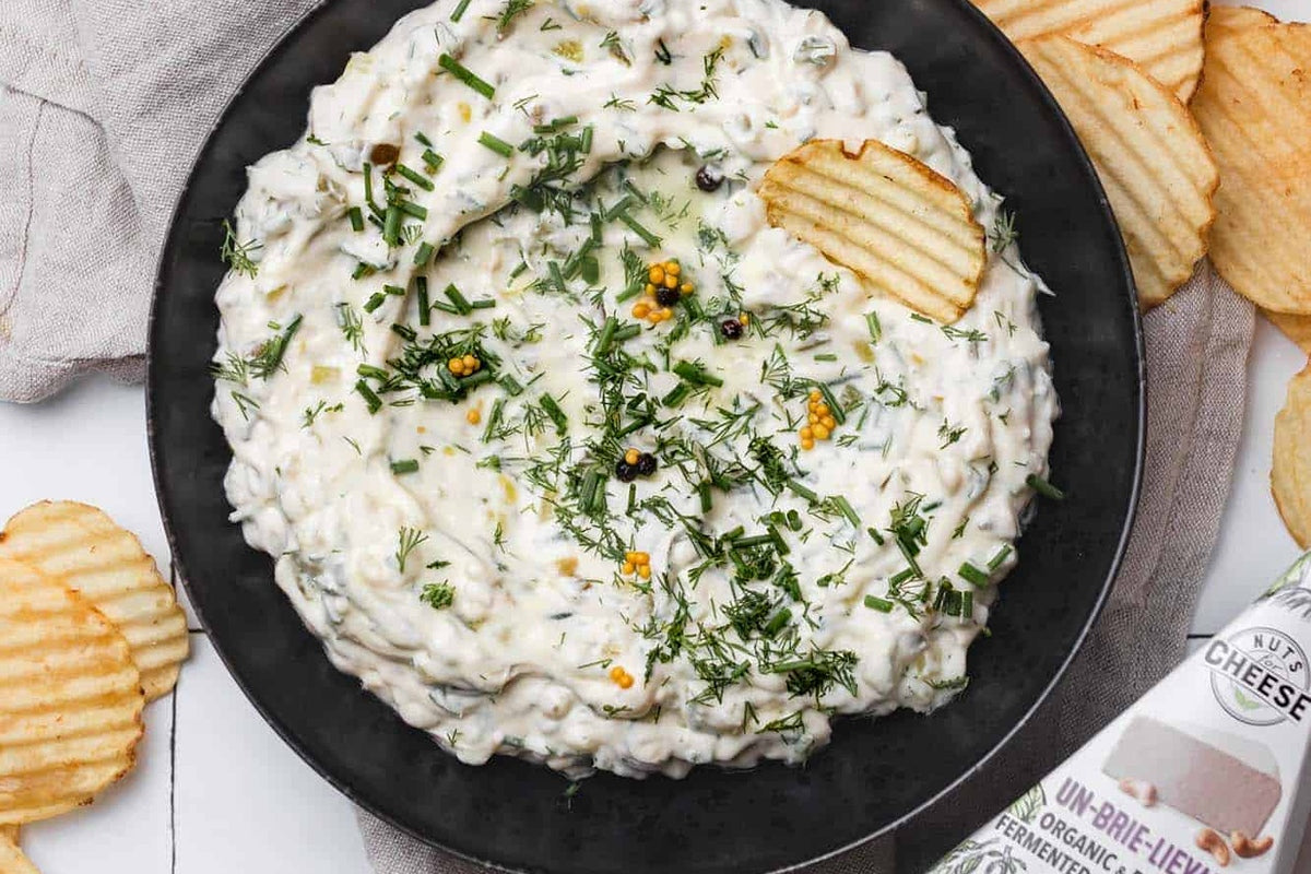 Bol de trempette de cornichons à l'aneth sans produits laitiers garni d'herbes fraîches et servi avec des chips à côté d'une boîte de brie sans produits laitiers....