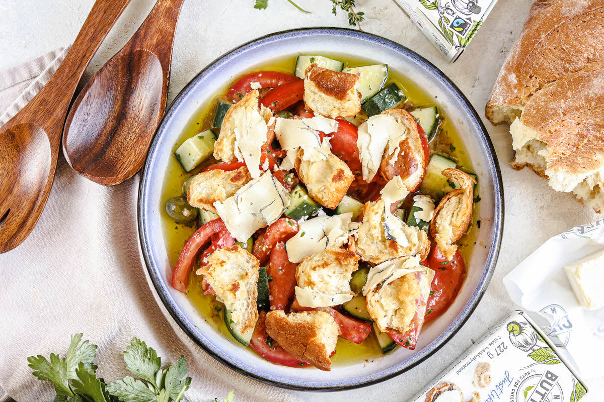 Bol de salade panzanella fraîche comprenant des tomates, des concombres, du pain croustillant et des morceaux de fromage à l'ail noir sans produits laitiers. Servi avec une miche de pain croustillant.