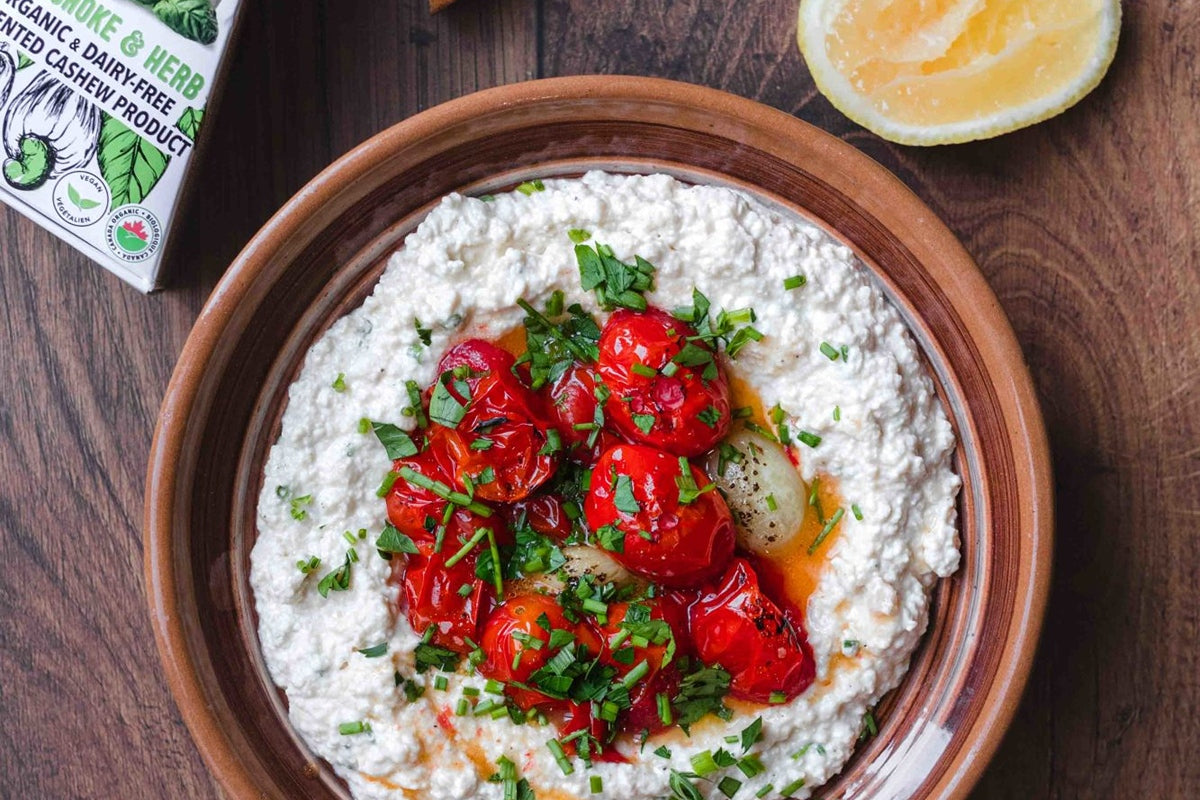 Bol préparé de trempette à la ricotta sans produits laitiers, garni de tomates rôties et de fines herbes. Disposé à côté d'une tranche de citron et d'une boîte de fromage d'artichauts et d'herbes sans produits laitiers.