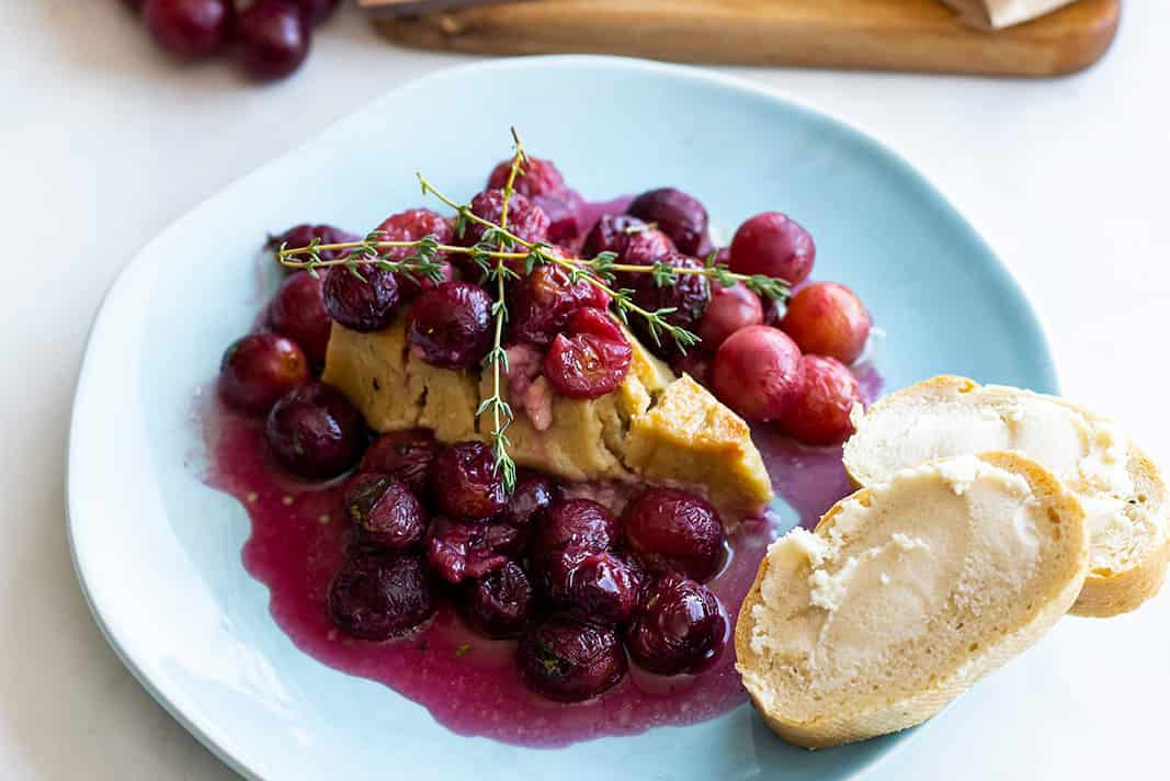 Une assiette de raisins rôtis sur un morceau de brie cuit au four sans produits laitiers, servie avec deux tranches de pain au levain.