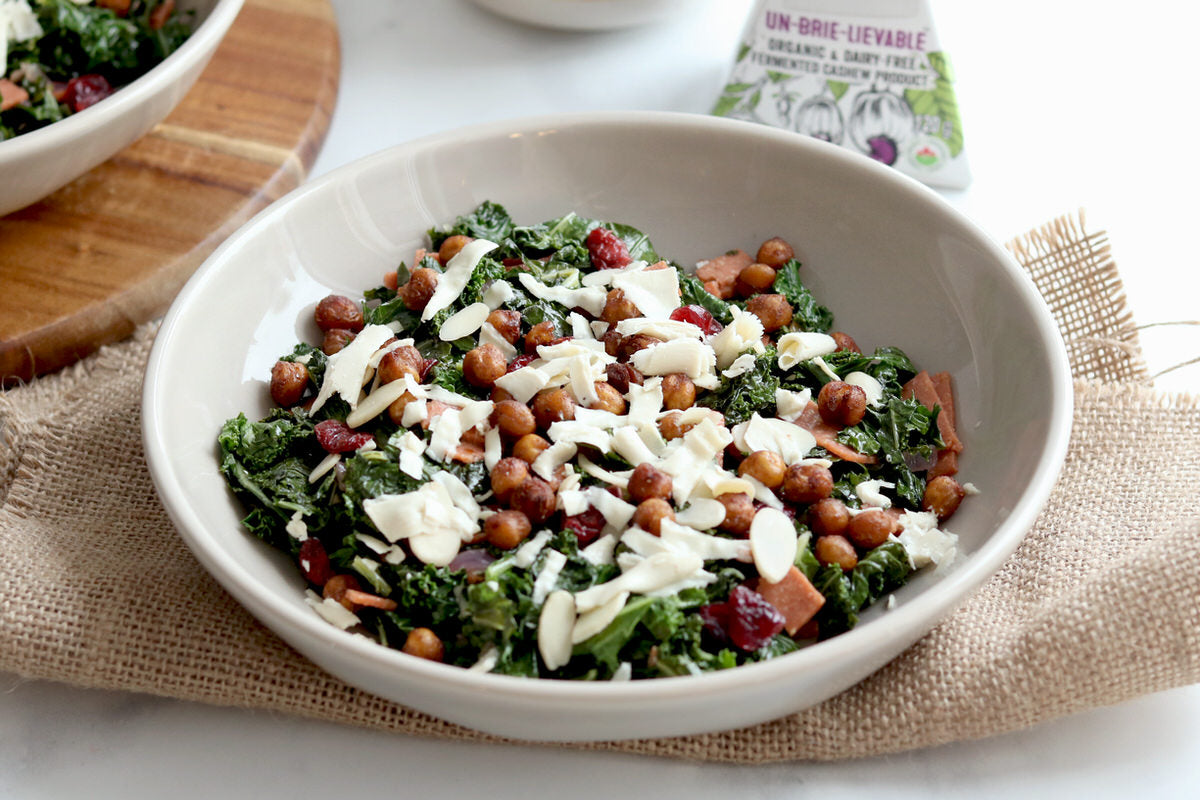 Grand bol de salade de chou frisé chaude garnie de pois chiches rôtis, de fromage sans laitage râpé et arrosée de vinaigrette au bacon végétalien. Servi à côté d'une boîte de fromage brie sans produits laitiers.