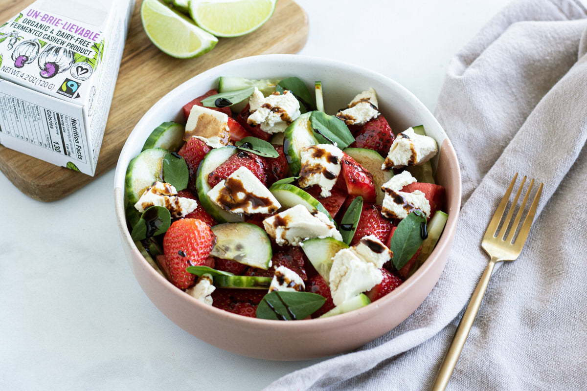 Bol de salade préparée comprenant des concombres, des fraises, de la pastèque et des morceaux de fromage sans lait, arrosé d'une vinaigrette balsamique. Servi à côté d'une boîte de brie sans produits laitiers et de quartiers de citron vert frais.