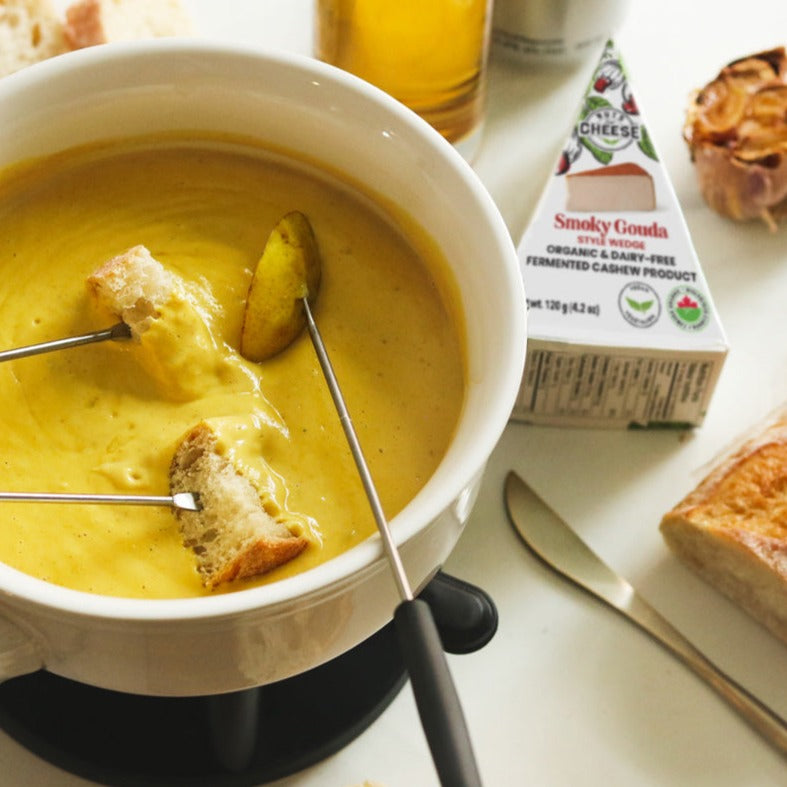 Un morceau de gouda fumé sans produits laitiers à côté d'un pot de fondue au fromage chaude sans produits laitiers avec des tranches de pain.