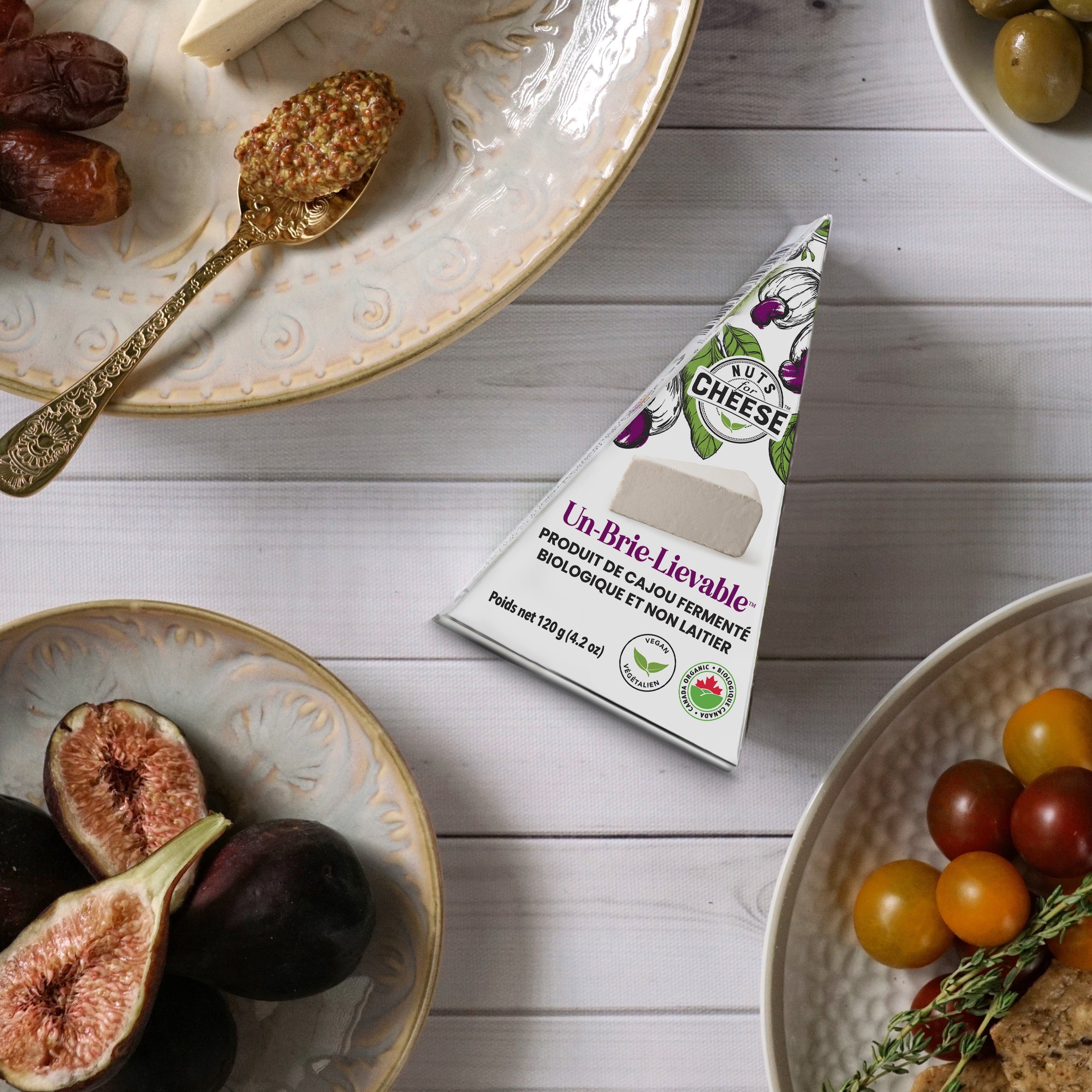 Nuts For Cheese™ Emballage de coins de fromage Brie Un-Brie-Lievable™ fermenté biologique et sans produits laitiers sur une table en bois blanc avec des olives, des figues et des craquelins.