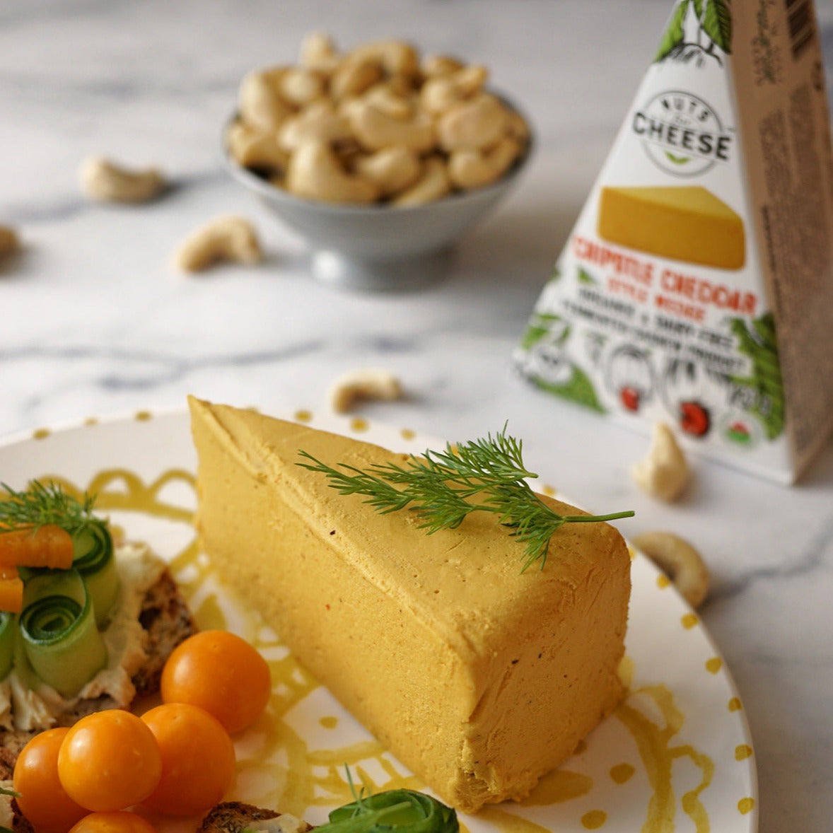 Nuts For Cheese™ Cheddar Chipotle fermenté biologique et sans produits laitiers en morceaux et emballé sur une assiette avec des morceaux de pain à la tomate et au concombre et des noix de cajou