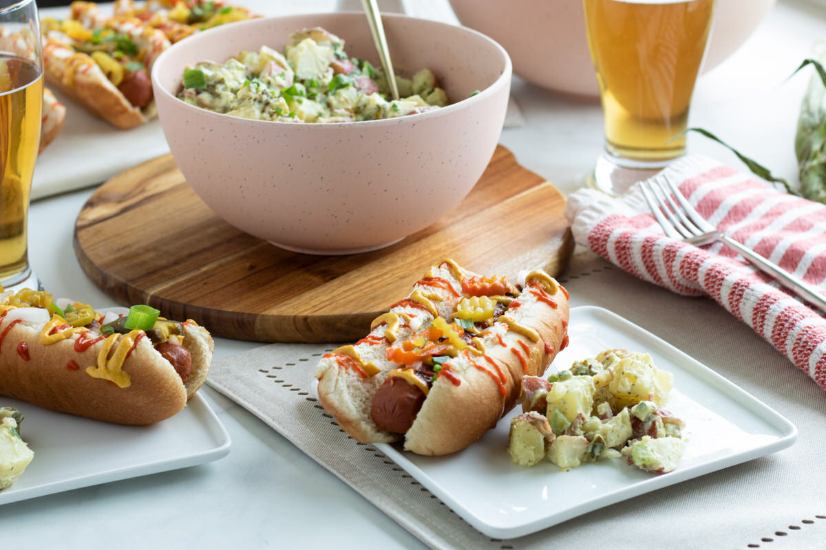 Deux assiettes contenant des hot-dogs végétaliens préparés, garnis de condiments et servis à côté d'une salade de pommes de terre végétalienne au fromage, avec un grand bol de salade de pommes de terre végétalienne en arrière-plan.