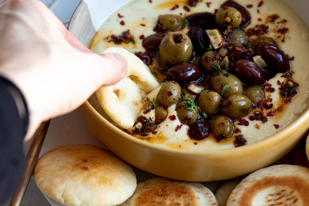 Une main trempe du pain dans un bol de trempette au brie fouetté sans produits laitiers, garni d'olives grillées. 
