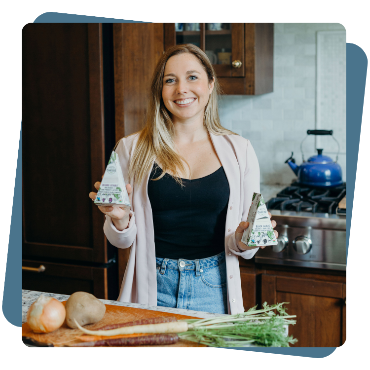 Margaret Coons, Nuts For Cheese™ Fondatrice, sourit en tenant des produits fromagers à base de plantes dans la cuisine avec des légumes sur le comptoir.